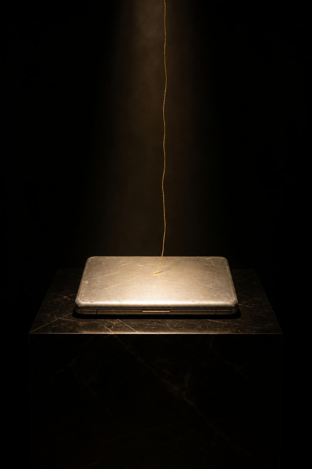 A scuffed silver laptop on a black marble plinth in a dark gallery, a thin gold thread descending onto its lid like a holy relic.